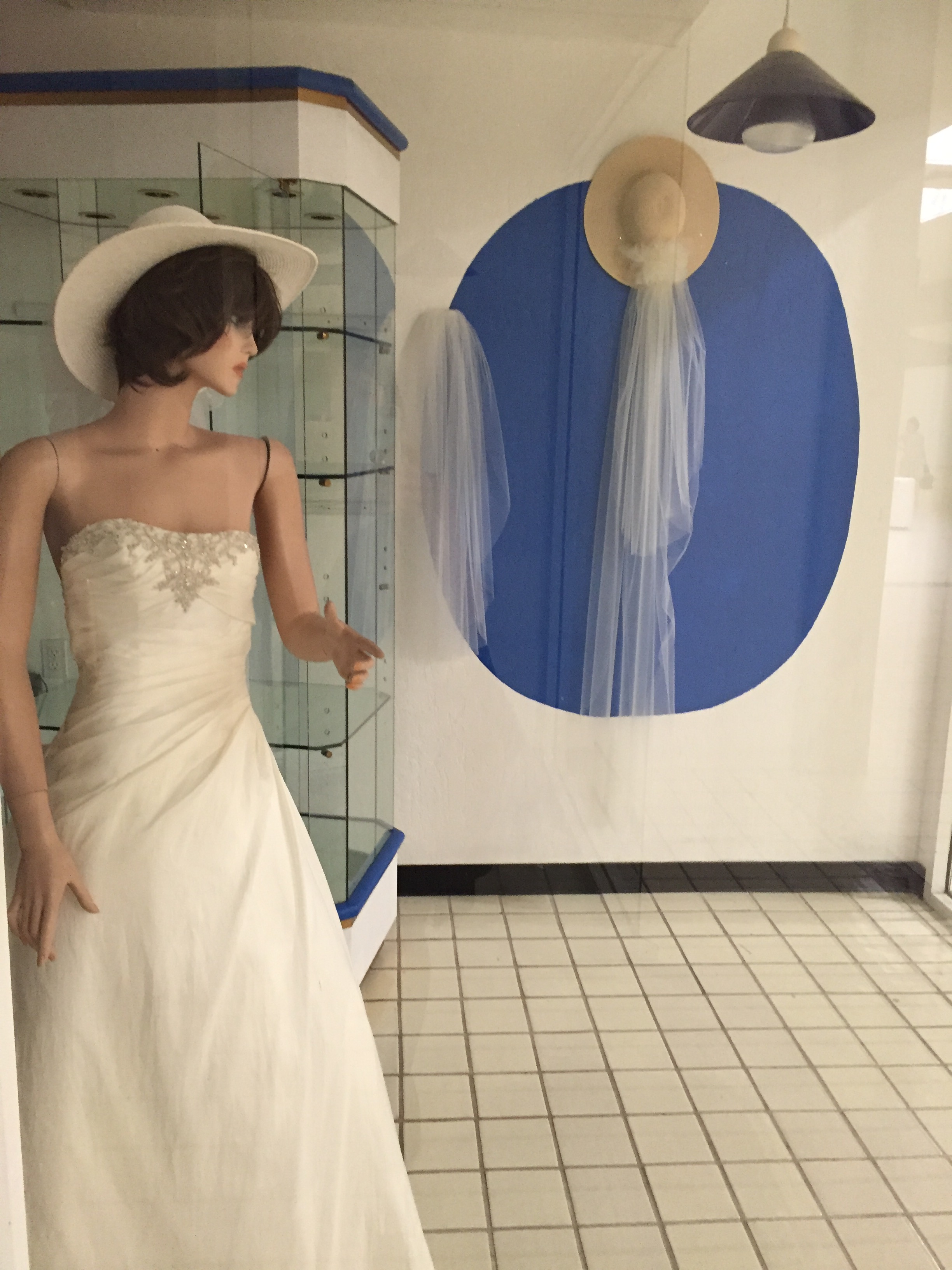 A mannequin in a white sun hat and a wedding gown points at the the viewer, looking away. On the wall behind the mannequin: a display of veils and sunhats.