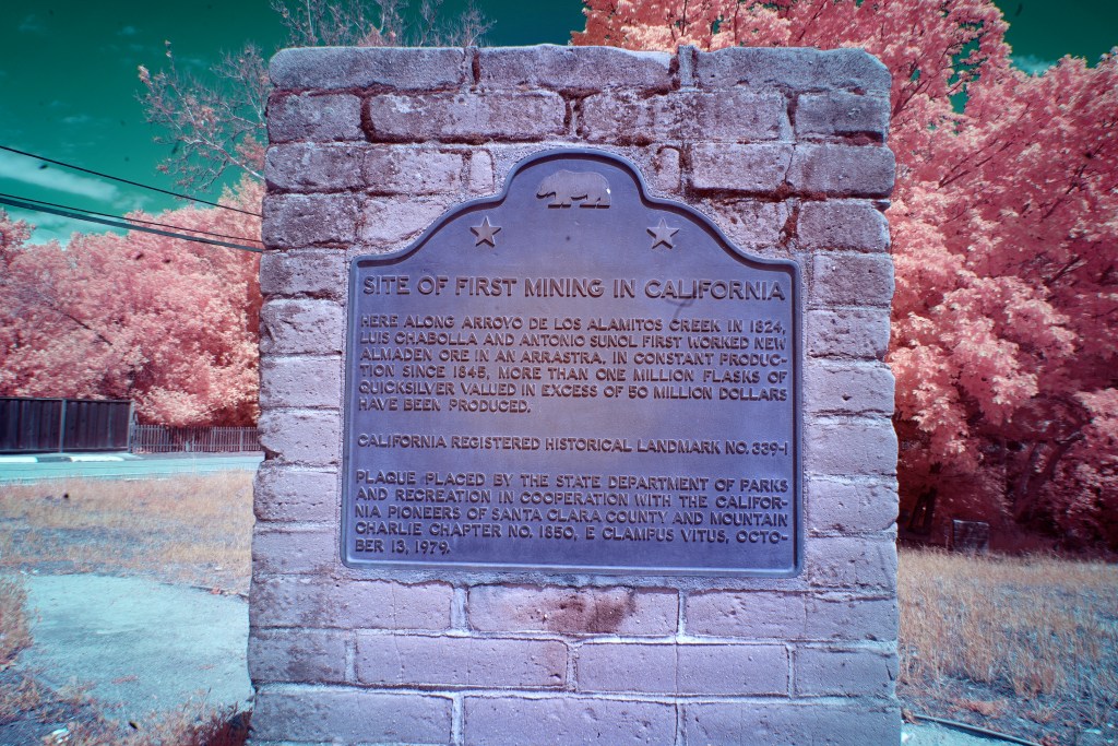 A color, infrared image shows a current brick monument at the site of the New Almaden Mines.  An aged plaque, with a california bear and two stars on the top reads, "SITE OF FIRST MINING IN CALIFORNIA: here along Arroyo De Los Alamitos Creek in 1824, Luis Chabolla and Antonio Sunol first worked New Almaden Ore in an Arrastra. in constant production since 1848, more than one million flasks of quicksilver valued in excess of 50 million dollars have been produced.  California registered historical landmark no. 399-1.  Plaque placed by the state department of parks and recreation in cooperation with the california pioneers of santa clara county and mountain charlie chapter no. 1850 E Clampus Vitus, October 13, 1979."  The brick of the monument is a peachy grey. The background shows some grass, a small picket fence on the left side, and some large, bushy trees.  The ground is a light teal and the sky is a dark teal.  The foilage is a light, peachy pink, which makes it look somewhat like cotton candy. 