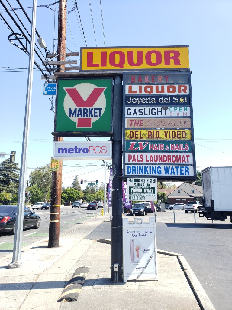 The strip mall's signage sits at the center of the image. Each store has a unique sign. The Y market sign is a large square, and a metro pcs sign sits beneath it. The top sign is a bright yellow and red "Liquor" sign, with "bakery", "liquor", "Joyeria del Sol", "Gaslight open 9 A.M", The (unreadable) factory", "Del Rio Video", "LV Hair & Nails", "Pals Laundromat", and "Drinking Water". There is a towing warning sign below and a Metro PCS foldable sign sitting on the ground. There are cars driving up and down white road in the background and a few trees are visible on the left side.