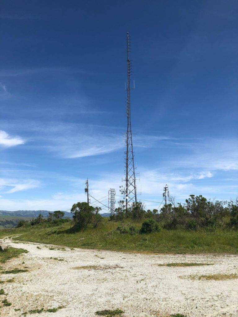 The top of Coyote Peak: a white gravel path surrounds a small patch of grass, atop which sits a huge antenna tower alongside more transmission towers and poles, in the background is a blue sky and a line of hills in the distance.