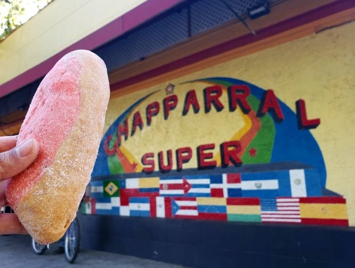 A brown hand holds a sugar coated pink and yellowish pan dulce. In the background is a painted sign that says "Chapparral Super" with flags from the Americas painted underneath. The building is yellow with orange and red trim, and there is a bicycle poking out from under the pan dulce on the left side.