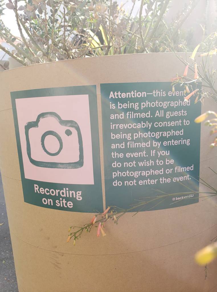 A short brown cylinder at the exit of Backyard.  It has two dark green signs on it.  In pink writing one says, "Attention - this event is being photographed and filmed.  All guests irrevocably consent to being photographed and filmed by entering the event.  If you do not wish to be photographed or filmed do not enter the event. @backyardSJ."  The second sign has a drawing of a camera with the words "Recording on site" underneath.  A wildflower pokes into the right side of the image. 