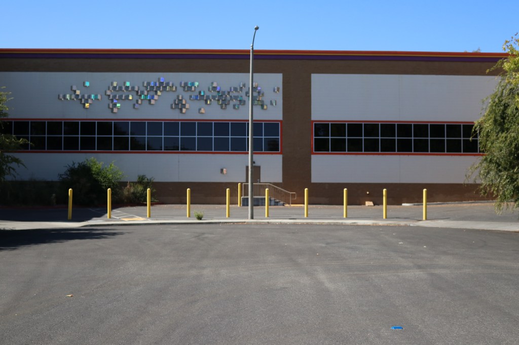 a side view of the Public Storage.  The foreground is a street cul-de-sac, with tree shadows on the left side.  In the center of the photo is some short polls that keep drivers from using this area as an entrance to Public Storage.  Behind the polls is the Public Storage itself, the roof is lined with a purple, yellow, and orange strip.  Some decorative metallic squares are on the upper left of the building, below are long, thin windows.  In the center of the building is a door with stairs going down to the cement.  There is a tree on either side of the image. 
