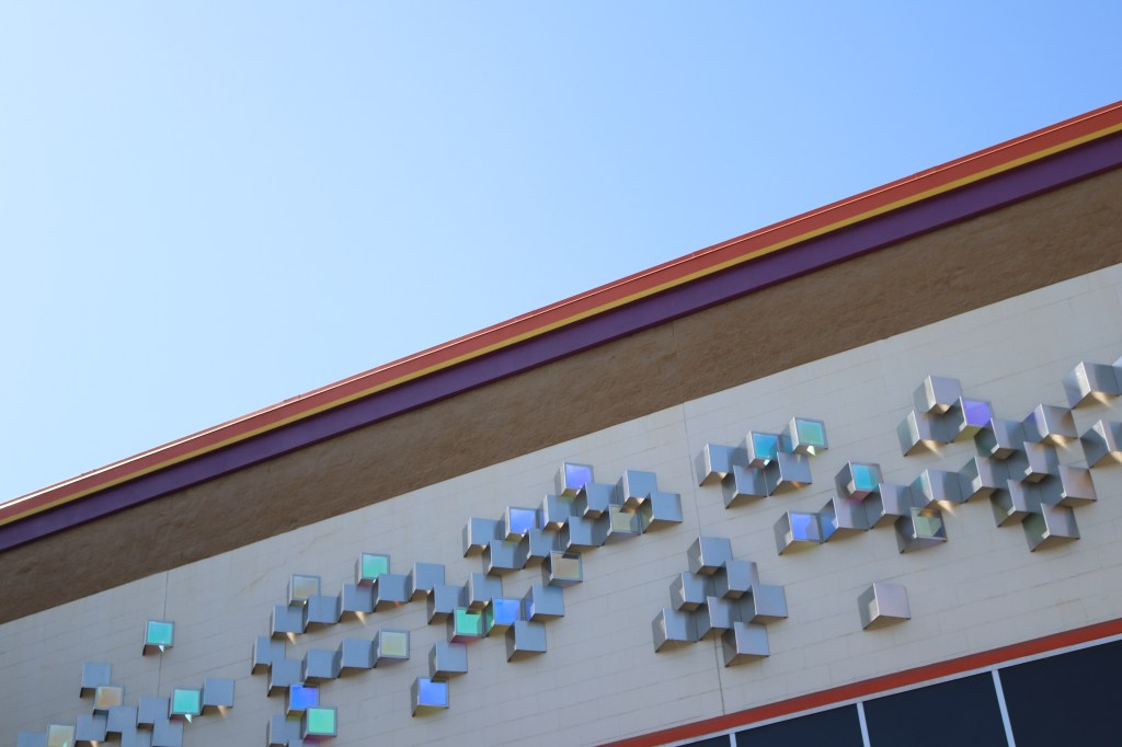 The photo is cut diagonally - in teh upper lefthand corner is the light blue sky.  Below is the top 10 feet of Public Storage.  An orange, yellow, and purple stripe line the roof.  below is a light brown strip, and below that are decorative metallic squares.  The windows peek out in the bottom righthand. 