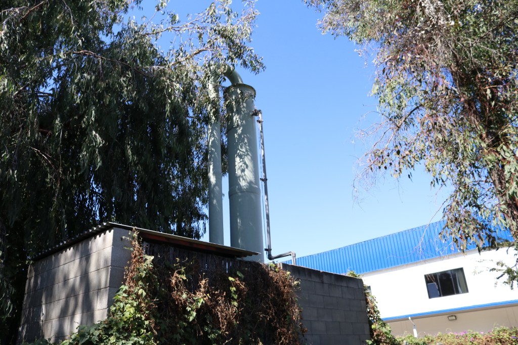 in the Public Storage forest, a mini-fortress houses a long, cylindrical metal contraption.  in the background is a blue and white building.  The fortress has green ivy growing on its side.  