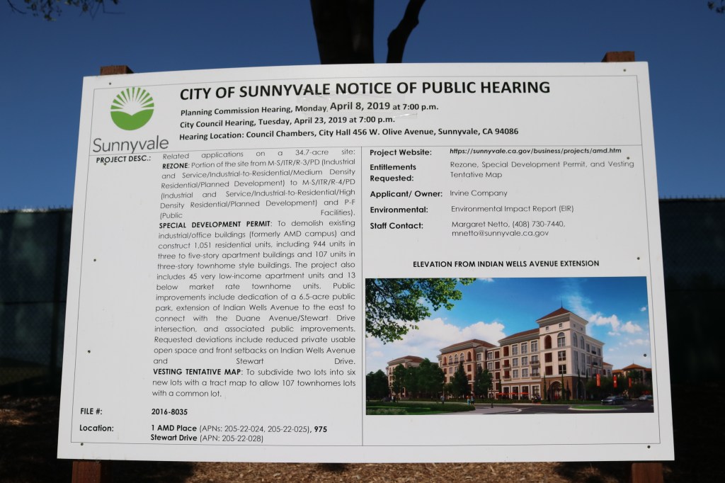A "City of Sunnyvale Notice of Public Hearing" sign sits in front of a green privacy screen and a blue sky.  On the sign is information from a public hearing that took place on April 8th, 2019.  The bottom right of the sign has an image of the future housing project that will be built in this location.  It is mixed use housing on a tree lined street. 