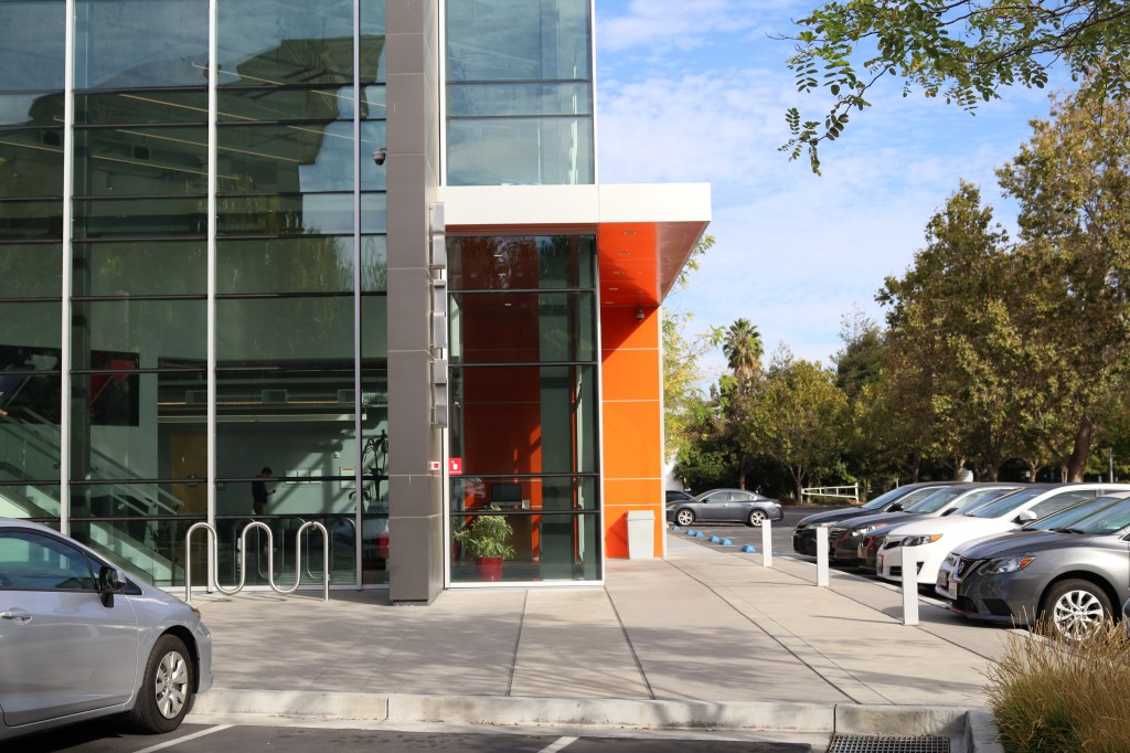 the entrance to small apple campus.  The building is all windows, and the entrance has a slick, bright orange overhang. The parking lot is lined with silver, white, and black cars.  The street is lined with trees and there is an empty bike rack outside.  A person stands inside the glass building, they are wearing a black shirt and looking down at their phone. 
