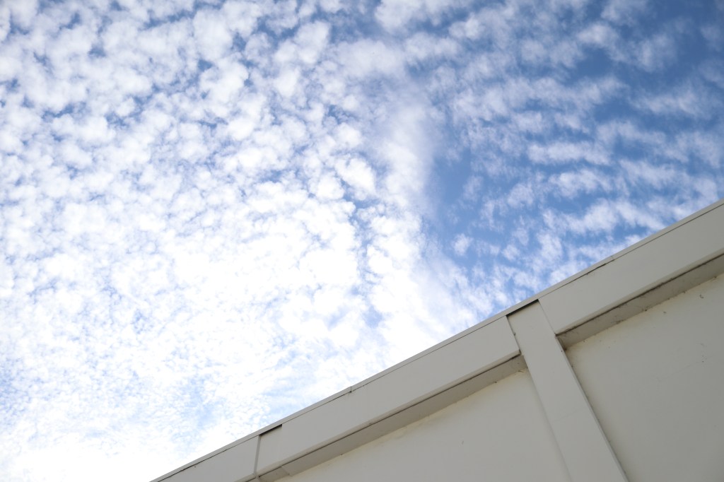 a diagonal photograph shows the upper part of a building roof.  A blue sky takes up the rest of the image, white clouds dot its surface.  