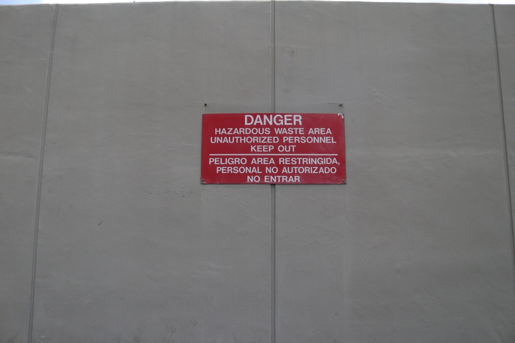 A red sign is on the side of a grey building, it reads, "Danger, hazardous waste area unauthorized personnel keep out.  Peligro area restringida personal no authrizado no entrar." 