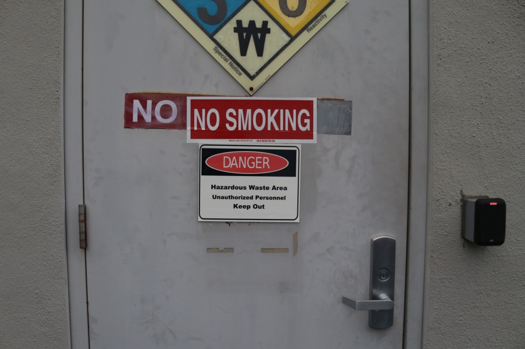 A 'no smoking' sign has been covered by a smaller 'no smoking sign', below it another sign reads, 'Danger, hazardous waste area unauthorized keep out."