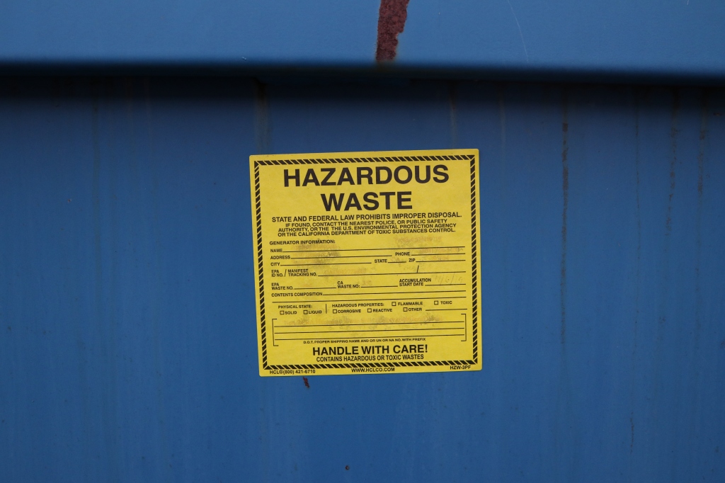 A blue dumpster has a large yellow sticker on its side.  Not all of the text is visible, and most are supposed to be filled in by someone.  In large letters at the top it reads, "Hazardous waste."  The bottom says, "Handle with care."