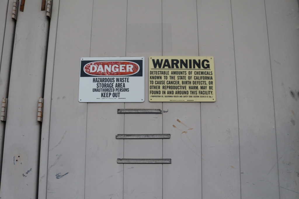 a white building has two warning sides on it.  One says, "Danger, hazardous waste storage area unauthorized persons keep out."  The other says, "Warning, detectible amounts of chemicals known to the state of california to cause cancer, birth defects, or other reproductive harm may be found in and around this facility."