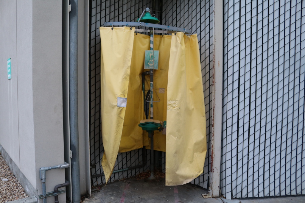 An eyewash station sits outside a grey building.  It looks like an old grey and green shower, a yellow shower curtain surrounds the eyewash station itself. 