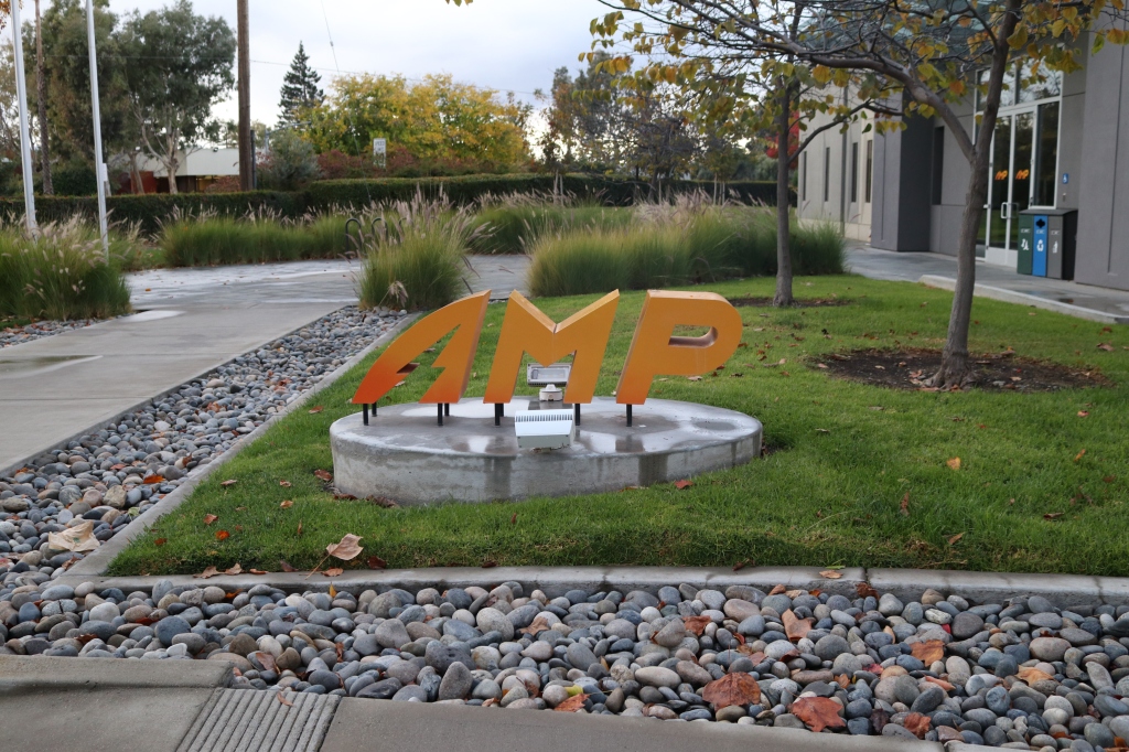 A set of large, orange, and slightly slanted letters spell out A.M.P.  They are surrounded by the manicured outside of a building.  The outside area is damp and grey, it's a rainy day and there are no folks to be seen, just some grass, rocks, and cement. 