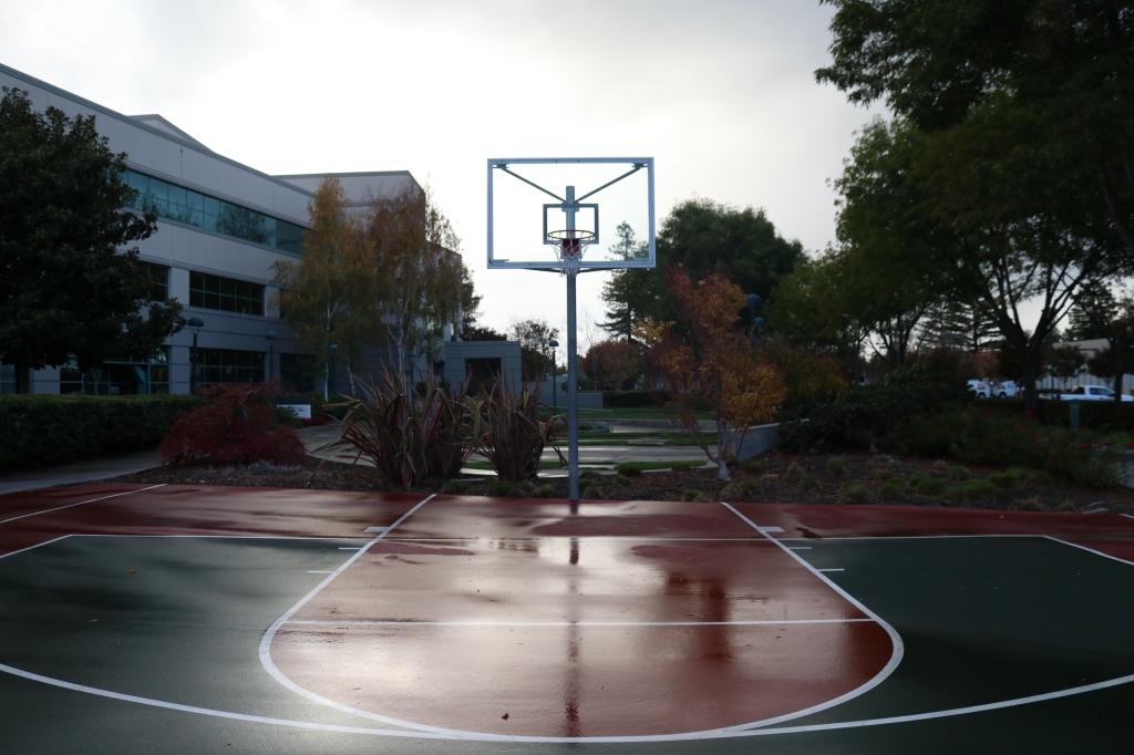A half basketball court is slick and shiny with rain.  The court is dark green and dark red, with a basketball hoop in the center of the image.  In the background are trees and a large office building. 