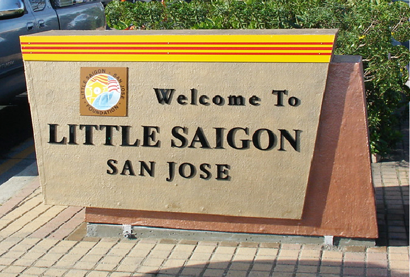 A sand colored sign, with an elongated Vietnamese flag across the top reads "Welcome to Little Saigon San Jose." A smaller logo sits on the left and it says "Little Saigon, San Jose Foundation." Behidn the sign is a green shrub and a portion of a blue Toyota Tacoma. 