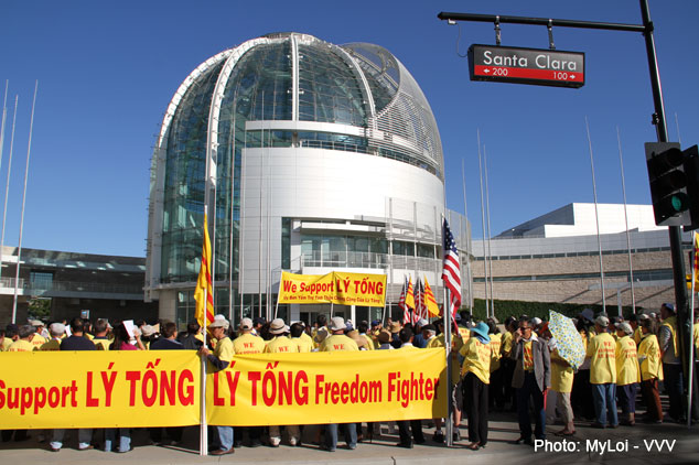 20 people stand in front of city hall, their backs to the camera.  Most are wearing a light yellow shirt.  City hall is in the background and there are various U.S and Vietnam flags in the foreground.  Two long banners read "We support Ly Tong." and another reads, "Support Ly Tong - Ly Tong Freedom Fighter."