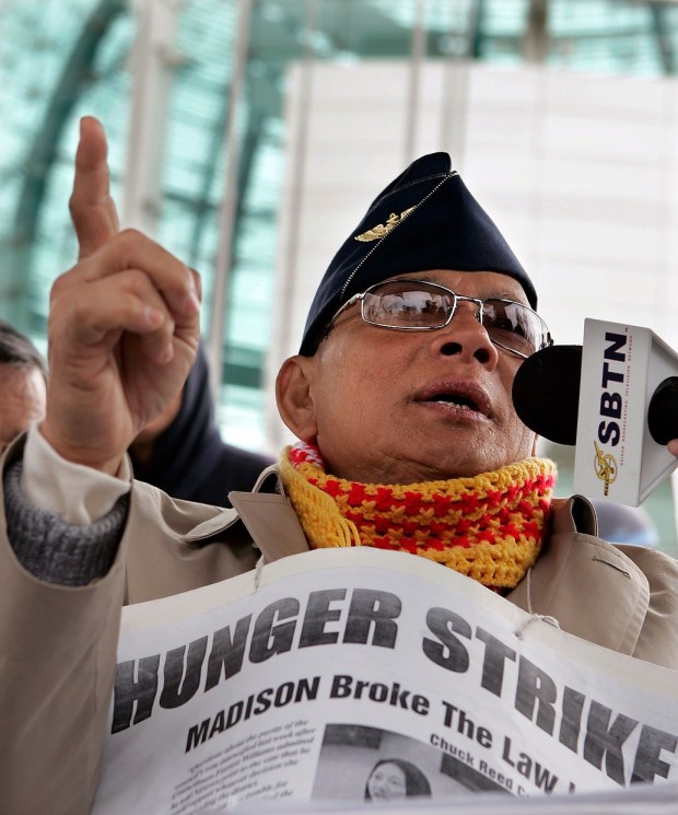A photograph of hunger striker, Ly Tong.  The image is looking up at Ly, he is speaking into a microphone and pointing up toward the sky with the index finger on his left hand.  He is wearing a military style hat, sunglasses, and a yellow and red scarf.  He has a enlarged newspaper clipping hanging from a string around his neck.  In large, bold letters on the top it reads, "Hunger Strike.", below "MADISON broke the law."